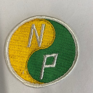 Pode incluir: Um remendo bordado branco, verde e amarelo com um símbolo yin yang. O símbolo tem as letras "N" e "P" dentro.