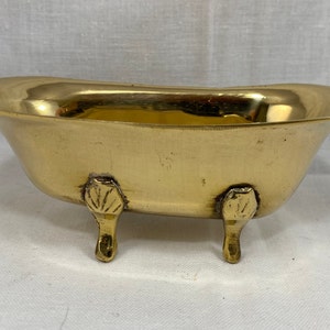 Puede incluir: Un pequeño plato de metal de color dorado en forma de bañera con tres patas decorativas. El plato está vacío y parece ser de latón.
