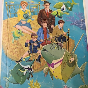 Może przedstawiać: Wzorowa układanka z ramą i tacą przedstawiająca postacie z filmu Disneya Bedknobs and Broomsticks. Układanka przedstawia scenę z łóżkiem unoszącym się na oceanie, otoczonym rybami i stworzeniami morskimi. Postacie obejmują kobietę w żółtej sukience, mężczyznę w garniturze, dziewczynkę z warkoczem, chłopca w niebieskiej koszuli i rybę z lornetką i cylindrem.