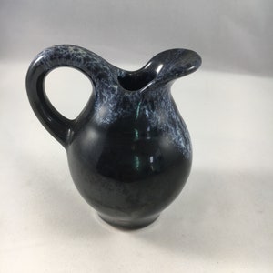 Puede incluir: Una pequeña jarra de cerámica negra con un esmalte blanco. La jarra tiene un cuerpo redondeado y un pico corto. El asa es curva y está unida al lado de la jarra.
