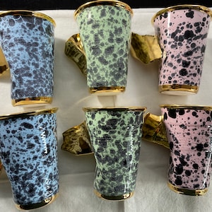 May include: A set of six ceramic cups featuring a speckled pattern in light blue, pale green, and soft pink. Each cup has a gold-colored rim and a distinctively shaped, metallic gold handle. The cups have a slightly irregular, textured surface.