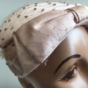 Puede incluir: Un sombrero de satén rosa claro con un lazo y un velo de red. El sombrero está decorado con pequeñas cuentas transparentes.
