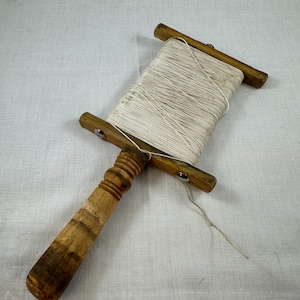 May include: A vintage wooden thread spool with a handle. The spool is filled with white thread and has a natural wood finish with darker brown accents. The handle is intricately carved.