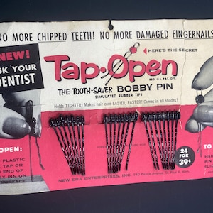 May include: Vintage package of "Tap-Open" bobby pins. The packaging features the text "No More Chipped Teeth!" and "The Tooth-Saver Bobby Pin." The package contains several black bobby pins. The packaging is red and white.