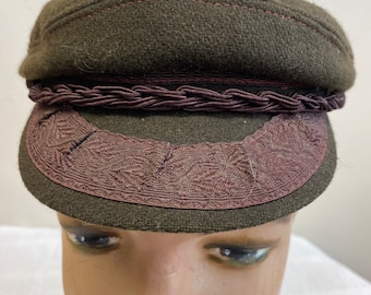 May include: A dark olive green cap with a flat top and a short, curved brim. The cap features a decorative braided band and a patterned trim around the brim. The stitching is a contrasting reddish-brown color.