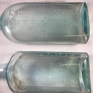 May include: Two vintage glass bottles with embossed text. The first bottle is clear and reads "LYDIA E. PINKHAM'S VEGETABLE COMPOUND". The second bottle is clear and reads "DR. PIERCE'S FAVORITE PRESCRIPTION".