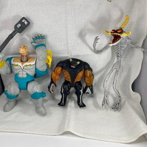 May include: Three action figures, one gray and blue with a weapon, one black with orange accents, and one gray with yellow accents.