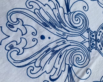 May include: Close-up of a white fabric with a repeating blue embroidered design. The design features stylized floral and scroll motifs, creating a pattern across the surface. The embroidery is detailed and adds texture to the fabric.