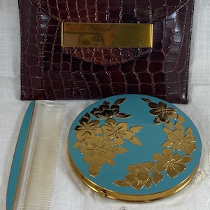 Op de afbeelding: Een vintage bruine clutch met krokodillenreliëf en een goudkleurige sluiting, een turquoise compactpoederdoosje met gouden bloemendetails en een bijpassende kam. De set wordt gepresenteerd op een wit oppervlak.
