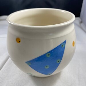 May include: A white ceramic pot with a blue, green, and yellow geometric design. The pot has a rounded shape and a wide opening.