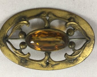 Op de afbeelding: Een ovale broche met een amberkleurige glazen steen gezet in een goudkleurig metalen frame. Het frame heeft een delicaat bloemmotief.
