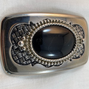 May include: A silver belt buckle with a black oval stone set in a decorative border. The buckle has a woven pattern around the stone.