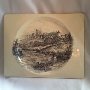 May include: Cream-coloured rectangular ashtray featuring a black and white illustration of Windsor Castle. The image is set within a circular indentation. The words "Windsor Castle" are printed below the image. The ashtray has a slightly raised edge.