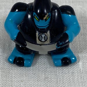 May include: A small, plastic action figure of a character from the Ben 10 series. The figure is primarily black and blue, with green eyes and a gray belt. The figure has a stylized design and is standing upright.