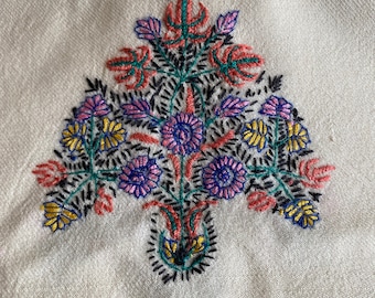May include: Cream-colored textile with a central embroidered floral design. The embroidery features a bouquet of flowers in shades of pink, blue, yellow, and green, with black and red accents. The design is symmetrical and detailed.