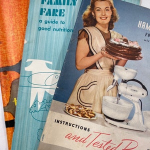 May include: Three vintage cookbooks stacked on top of each other. The top book features a woman holding a chocolate cake and a stand mixer. The book title is "Instructions and Tested Recipes". The middle book is blue and has the title "Family Fare: A Guide to Good Nutrition". The bottom book is orange.