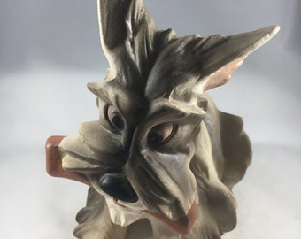 Peut inclure: Figurine de chien en céramique au design stylisé et caricatural. Le chien est principalement gris avec des accents beiges, un nez noir et une bouche rouge. Les oreilles sont grandes et pointues, et le chien semble tenir quelque chose dans sa gueule.