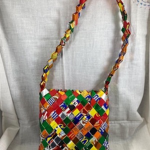 May include: A colorful, woven shoulder bag made from recycled materials. The bag features a patchwork design with various colors and patterns, including text from packaging. The strap is also made from the same material.