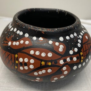 May include: A small, black ceramic pot with a brown and white painted design of an animal. The pot has a round body and a wide opening.