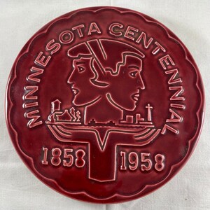 Puede incluir: Un plato de cerámica granate con borde festoneado. El plato presenta las palabras "MINNESOTA CENTENNIAL" alrededor del borde, con los años 1858 y 1958. El diseño central incluye dos perfiles y un paisaje.