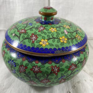 Op de afbeelding: Ronde, dekselbare sieradendoos met een groene achtergrond en bloemenmotief. De doos heeft een blauwe rand en gouden afwerking. Het deksel heeft een kleine knop bovenop. De bloemen zijn rood, geel en blauw.