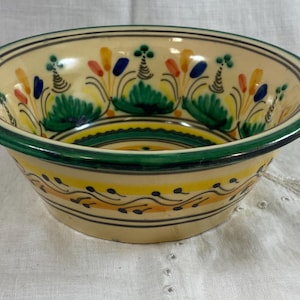 May include: A ceramic bowl with a cream-colored base and a green rim. The bowl is decorated with colorful floral patterns in green, yellow, orange, and blue. The bowl's exterior features a yellow band with black dots.