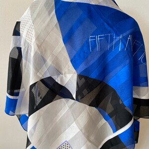 May include: A silk scarf with a geometric pattern in blue, white, black, and gray. The scarf has the words "FIFTH AVE" in white script. The scarf is draped, showing the pattern and colors.
