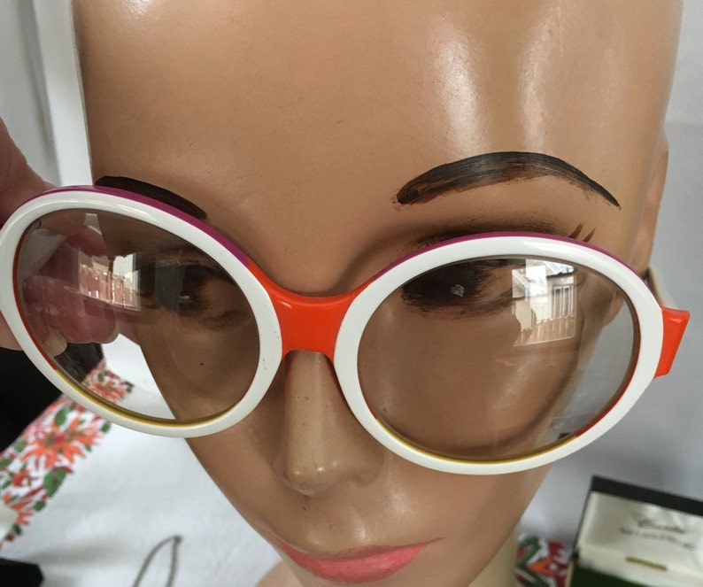 May include: White round sunglasses with a pink and orange trim. The sunglasses are on a mannequin head.