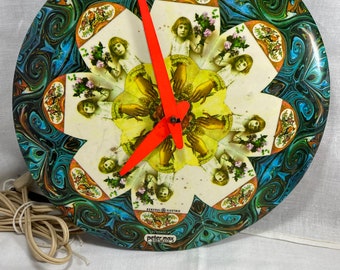 May include: Vintage General Electric clock with a Peter Max design. The clock face features a floral pattern with a swirling blue, green, and brown design. The center has a flower shape with images of children holding flowers. The clock hands are bright orange.