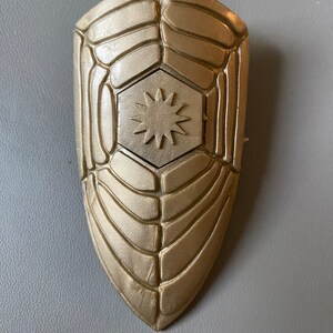 May include: A gold-colored shield with a hexagonal design and a star in the center. The shield has a textured surface.