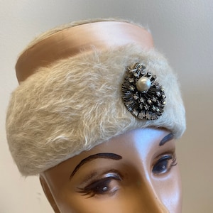 May include: A white faux fur hat with a pink satin band and a silver and rhinestone brooch with a pearl in the center.