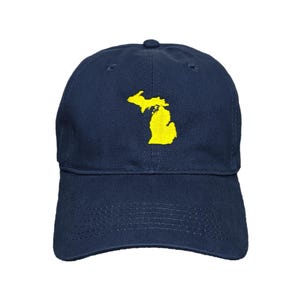 May include: A navy blue baseball cap featuring a yellow embroidered outline of the state of Michigan. The cap has a curved peak and an adjustable strap. The hat is made of a soft, durable fabric.