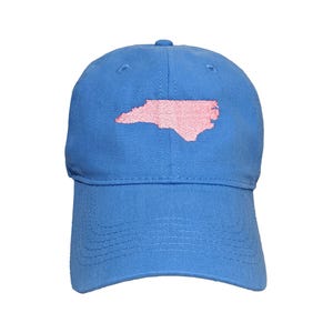 May include: A blue baseball cap with a pink embroidered outline of the state of North Carolina. The cap has a curved brim and an adjustable strap at the back. The hat is made of a soft, durable fabric.