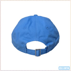 May include: A light blue baseball cap, viewed from the back, with a curved brim and adjustable strap. The cap has a metal buckle and a small button on top. The fabric appears to be a sturdy cotton or similar material.