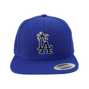 May include: A royal blue snapback cap featuring an embroidered white and black palm tree and the letters LA on the front panel. The flat brim includes a circular sticker with the text The Authentic Yupoong Snapback Yupoong Since 1974.