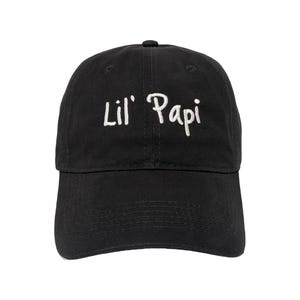 May include: A black baseball cap featuring the text Lil' Papi embroidered in white thread across the front panel. The cap has a structured crown, a curved brim, and a button at the top centre, providing a clean and simple design for headwear.