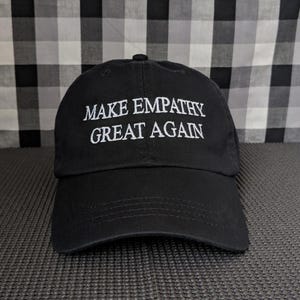 Peut inclure: Casquette de baseball noire avec l'inscription blanche brodée "MAKE EMPATHY GREAT AGAIN". La casquette a une visière incurvée et un bouton sur le dessus. Le fond est un motif à carreaux noir et blanc.