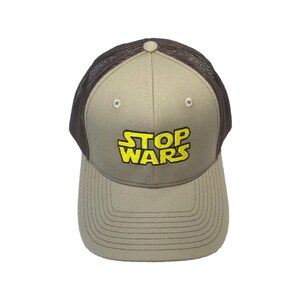 May include: A beige baseball cap with a brown mesh back and a curved brim. The front panel features the text STOP WARS embroidered in yellow with a black outline. The cap includes a matching beige button on the top and stitched detailing.