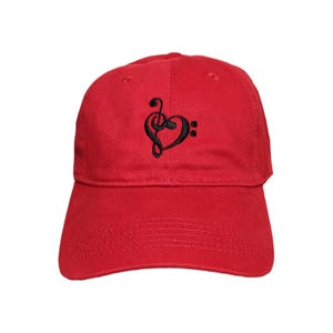 May include: A bright red baseball cap featuring a black embroidered design on the front panel. The embroidery depicts a musical heart shape formed by combining a treble clef and a bass clef symbol, centered above the curved brim of the cap.
