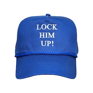 May include: A blue trucker hat with the text "LOCK HIM UP!" in white block letters. The hat has a mesh back and a curved brim. A blue rope detail accents the brim.