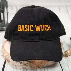 May include: A black baseball cap featuring the text BASIC WITCH embroidered in orange thread on the front panel. The cap has a curved brim and a structured crown, showcasing a classic six-panel design with a button at the top center.