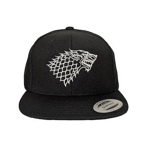 May include: A black snapback cap featuring a white embroidered direwolf head logo on the front panel. The hat has a flat brim and includes a circular silver and black sticker on the right side of the brim that reads The Authentic Snapback.