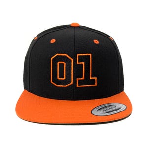 May include: Black and orange baseball cap featuring the number "01". The cap has a flat peak and a snapback fastening. The design includes orange stitching and a contrasting orange peak. A classic, stylish accessory.