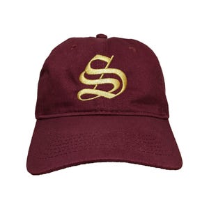 Puede incluir: Gorra de béisbol granate con una letra "S" dorada bordada en la parte delantera. La gorra tiene una visera curva y una correa ajustable en la parte trasera. El sombrero está hecho de una tela suave y duradera.