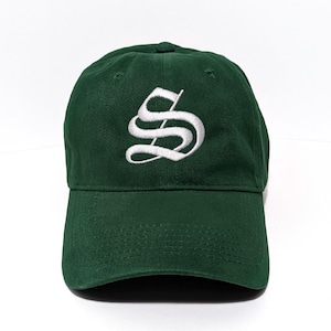 May include: A dark forest green baseball cap featuring a white embroidered letter S in a gothic-style font centered on the front panel. The cap has a curved brim, a structured crown with eyelets, and a matching green button on the top center.