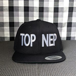 May include: A black snapback cap featuring the white embroidered text TOP NEP on the front panel. The hat has a flat brim with a circular sticker on the right side and a structured crown with ventilation eyelets on the top panels.