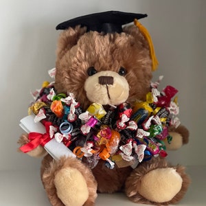 May include: A brown teddy bear wearing a black graduation cap with a yellow tassel. The bear is holding a white scroll with a red ribbon and is surrounded by a colorful assortment of candy.