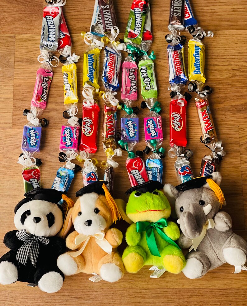 Op de afbeelding: Vijf pluche speelgoed met een afstudeerthema met afstudeerhoeden en kwastjes. Elk speelgoed houdt een snoepketting vast gemaakt van verschillende chocolade- en snoeprepen, waaronder Airheads, Butterfinger, Kit Kat, Twix en Laffy Taffy.