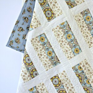 May include: A quilted blanket featuring a repeating pattern of rectangular blocks with cream, blue, and yellow floral and geometric motifs. The quilt is bordered by a light blue fabric with a small dark floral print and has white stitched accents.