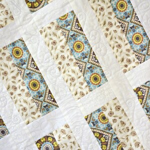May include: A quilted fabric runner featuring a patchwork design of cream, light blue, and yellow floral and geometric patterns. The textile includes white fabric strips with intricate, swirling machine-stitched quilting details throughout.